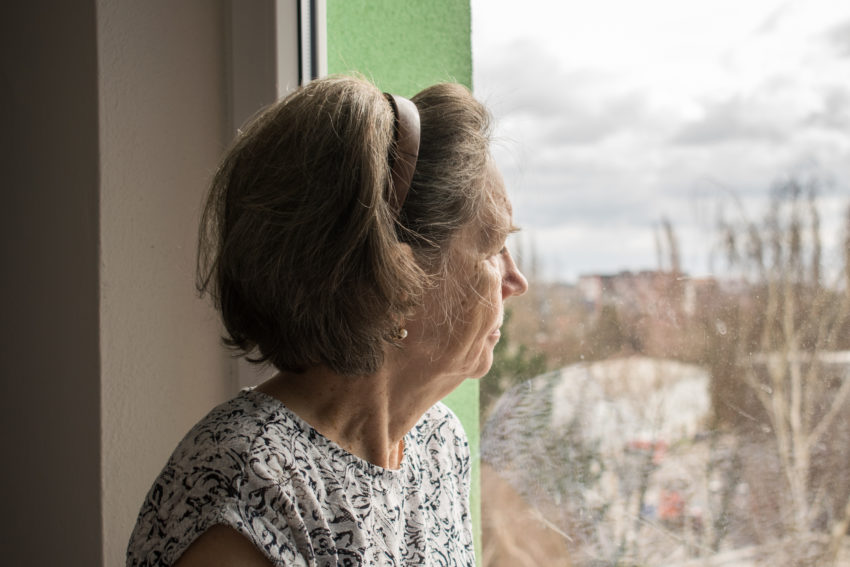 An older woman looks out of a window at overcast skies, she's wearing a white t-shirt decorated with black patterns.