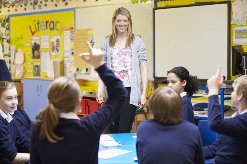 Blonde female teacher wearing a grey cardigan and floral top is stood at the front of a class, teaching modern foreign languages to young students. The children are wearing navy sweatshirts and white polo shirts, sitting round a table. All 