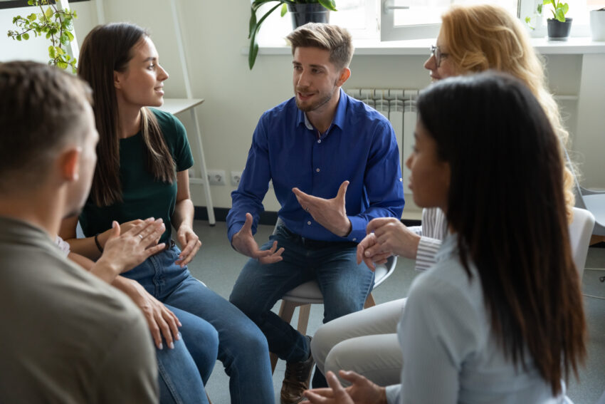 A group of employees sat on chairs in a circle sharing ideas and discussing mental health experiences, supporting each other in the work place. There are two white men, one in centre frame and the other with his back to the camera. Three women, one is mixed race and the other two are white.